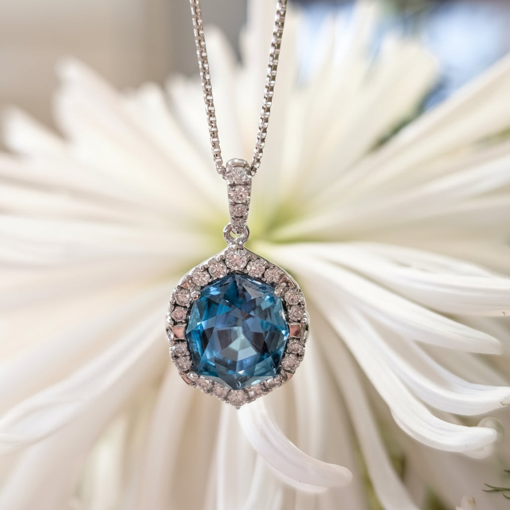 A silver-toned necklace with a heart-shaped pendant featuring a large faceted blue gemstone surrounded by small clear stones, displayed against a blurred white flower background.