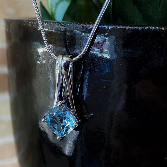 Photo of "V" Pendant with Blue Topaz Accent in a decorative scene, designed by , available at Plumb Gold & Plumb Silver