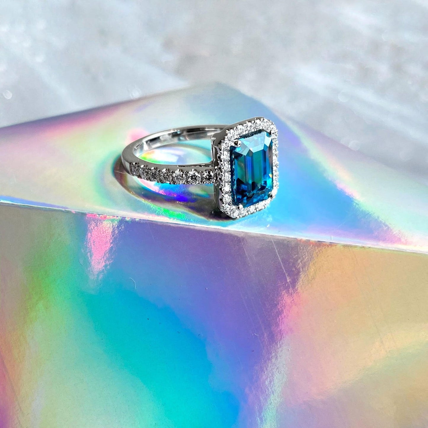 Photo of Zoma Color Emerald Cut Blue Zircon Ring with Diamond Halo in a decorative scene, designed by , available at Plumb Gold & Plumb Silver