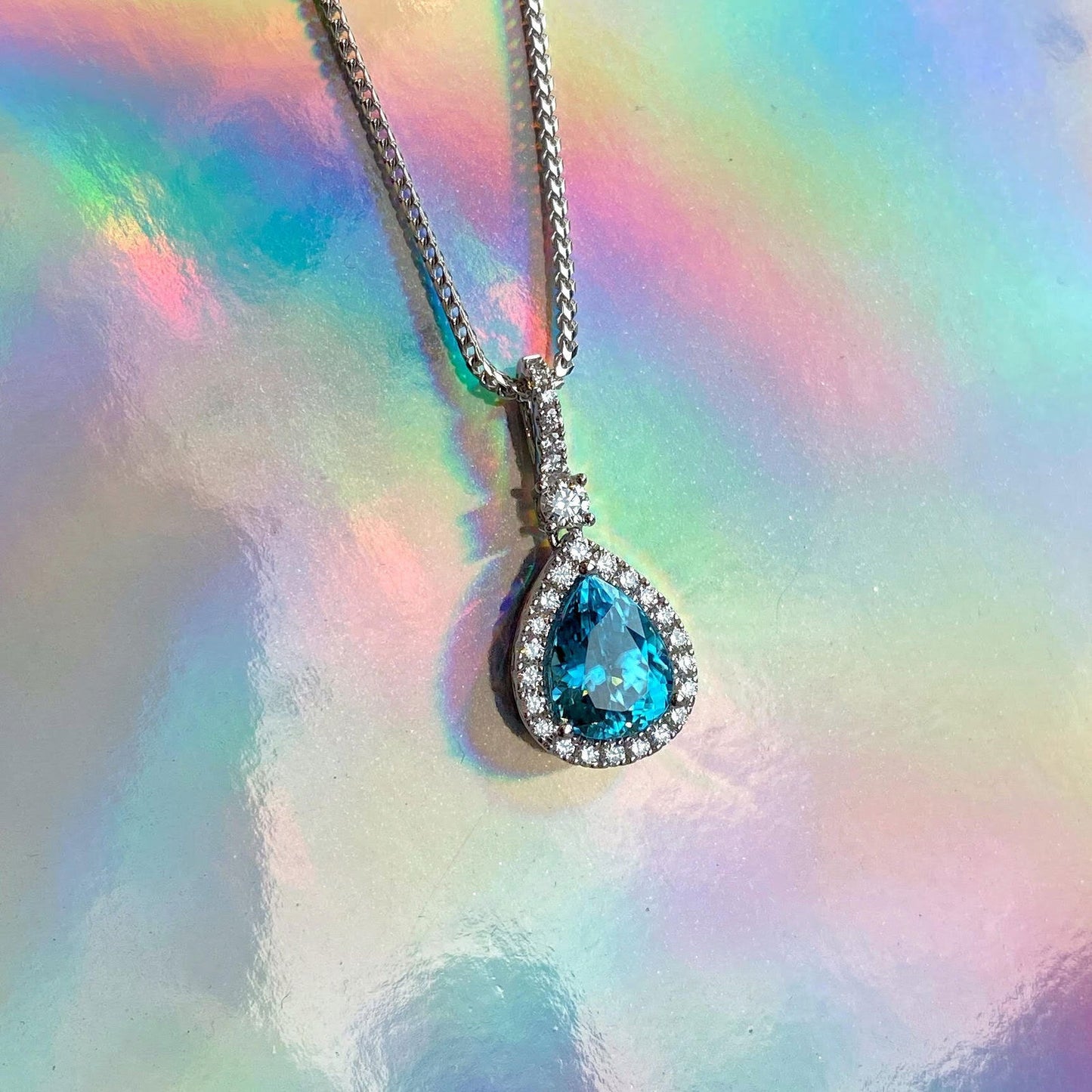 Photo of Zoma Color Pear Shaped Blue Zircon &amp; Diamond Pendant in a decorative scene, designed by , available at Plumb Gold & Plumb Silver
