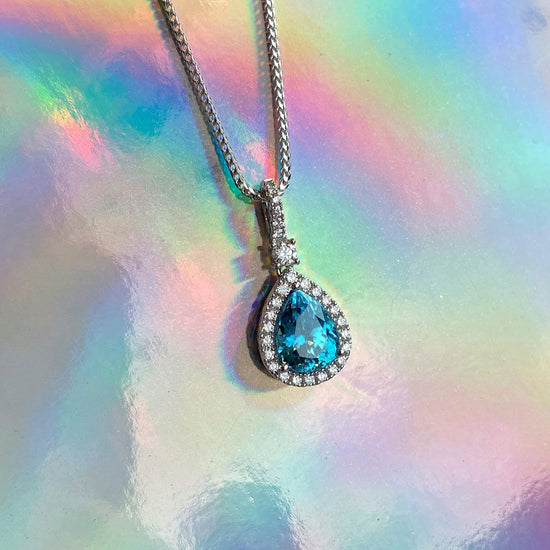Photo of Zoma Color Pear Shaped Blue Zircon &amp; Diamond Pendant in a decorative scene, designed by , available at Plumb Gold & Plumb Silver